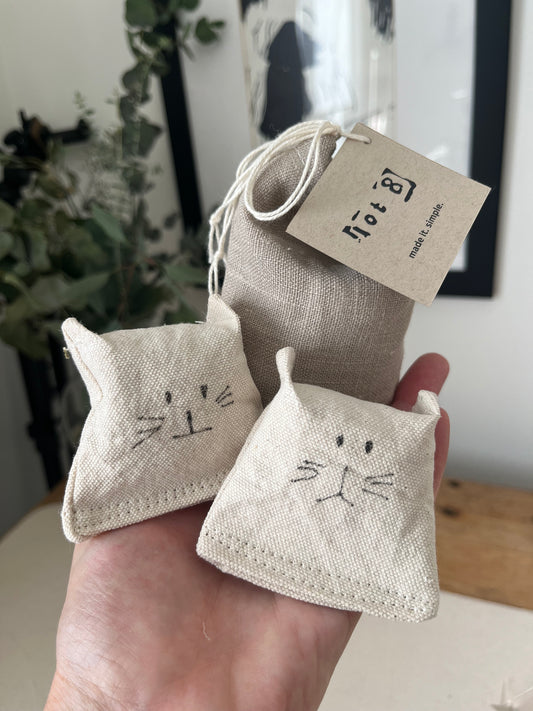 2 all-natural cat toys with adorable drawn cat faces rest in the palm of a hand alongside a linen pouch of cat grass seed. A framed image of the silhouette of a woman and a vase of eucalyptus is in the background.