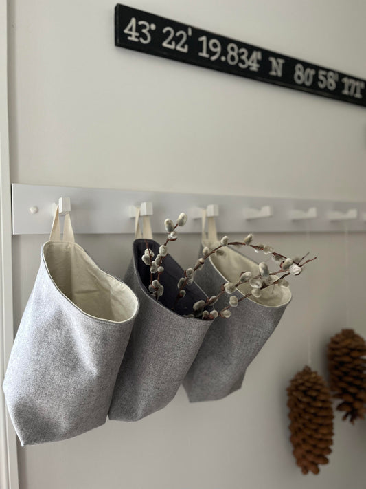 3 all purpose sacks hang from a white hanging rail in a foyer. One of the sacks is filled with pussy willows. A couple of pinecones are blurred in the distance hanging from the same rail.