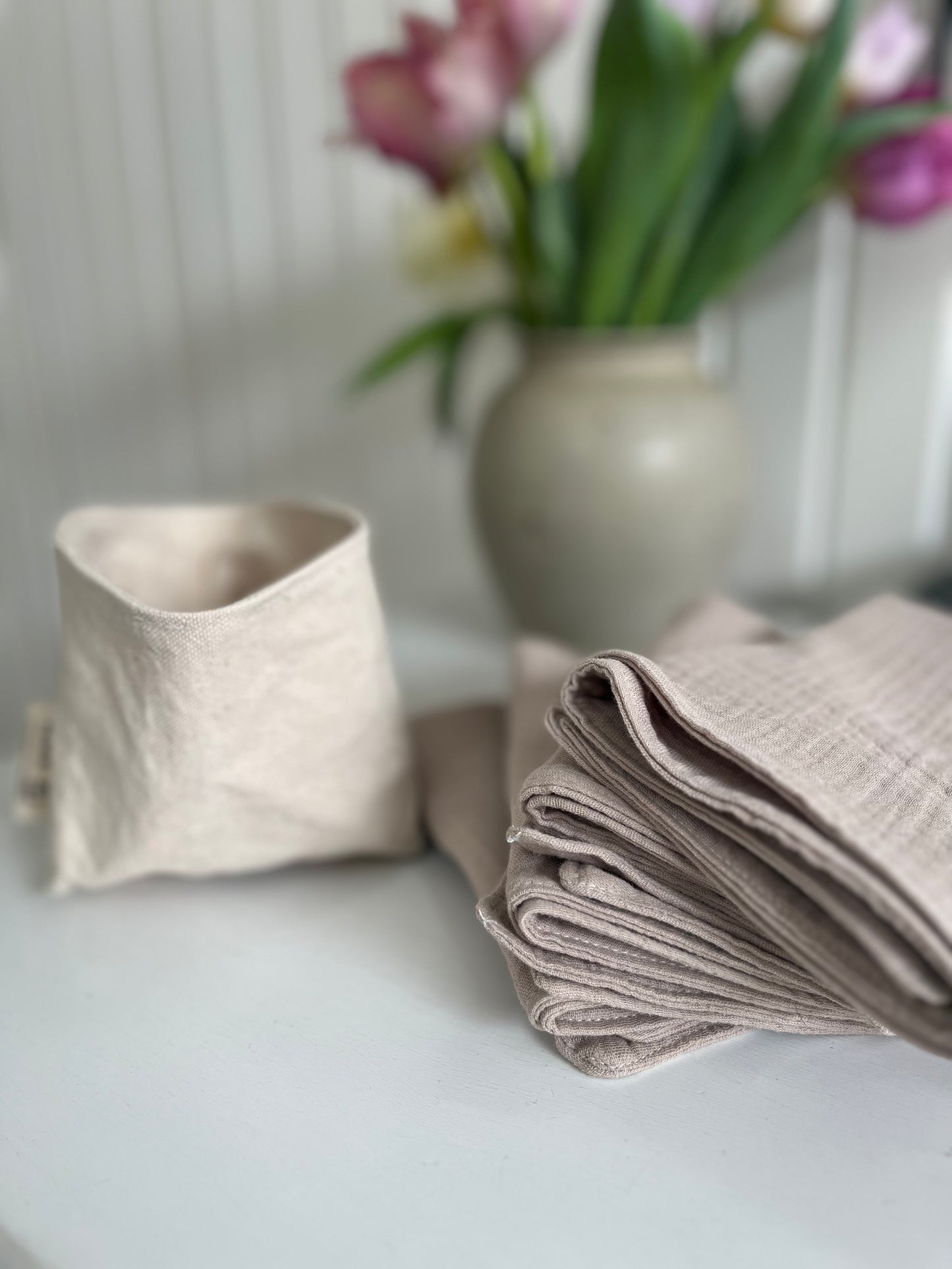 4 organic cotton facial cleansing cloths lay in a stack beside an empty fabric organizer called The Bucket. A vase of pink tulips is blurred in the background.