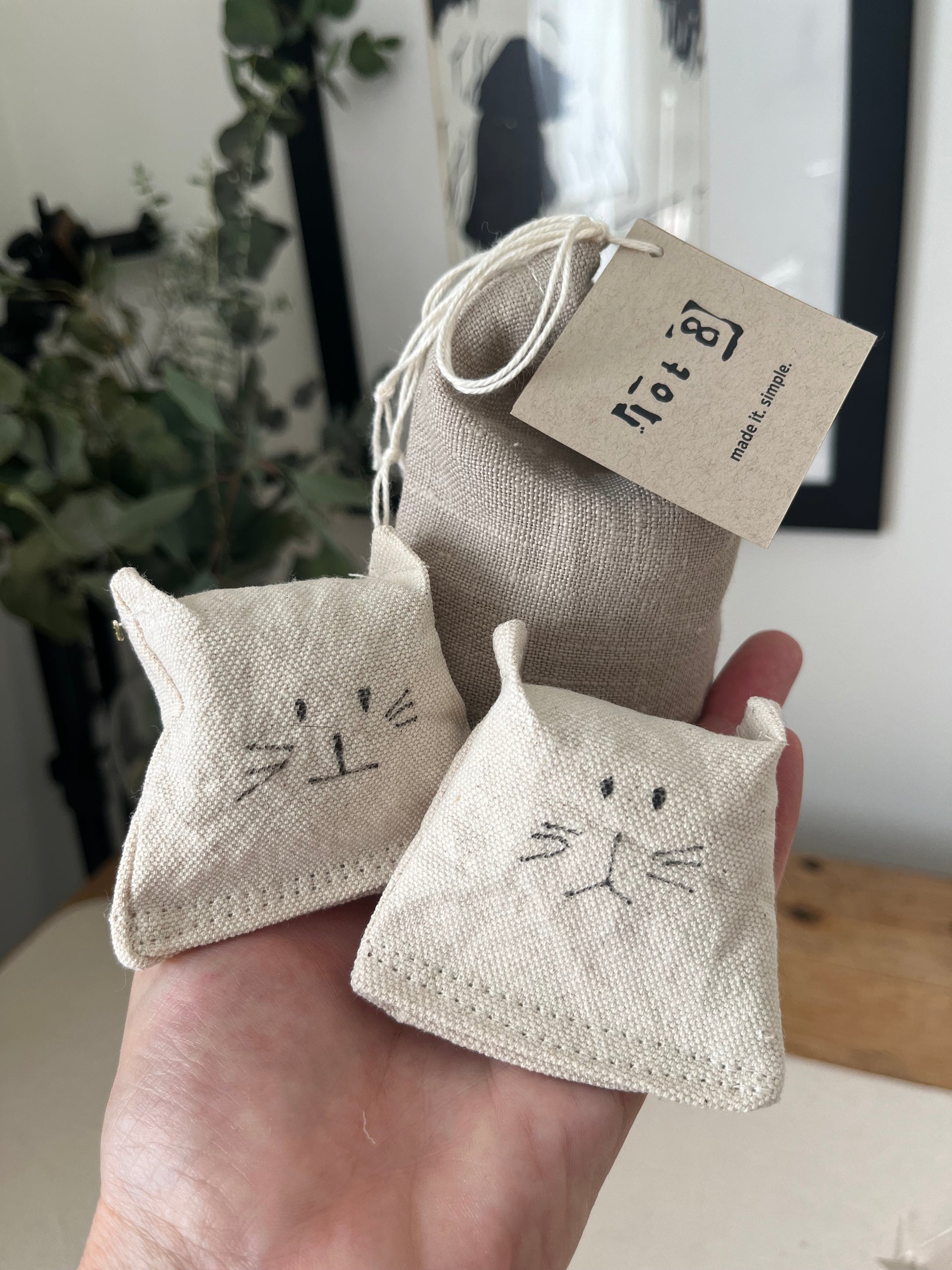 2 all-natural cat toys with adorable drawn cat faces rest in the palm of a hand alongside a linen pouch of cat grass seed. A framed image of the silhouette of a woman and a vase of eucalyptus is in the background. 