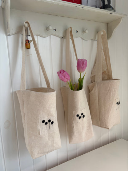 3 cotton canvas posy totes hang from a peg rail. Pink tulips poke their heads from the centre tote. Each tote has a handmade appliqued stamp of billy buttons in black on canvas.