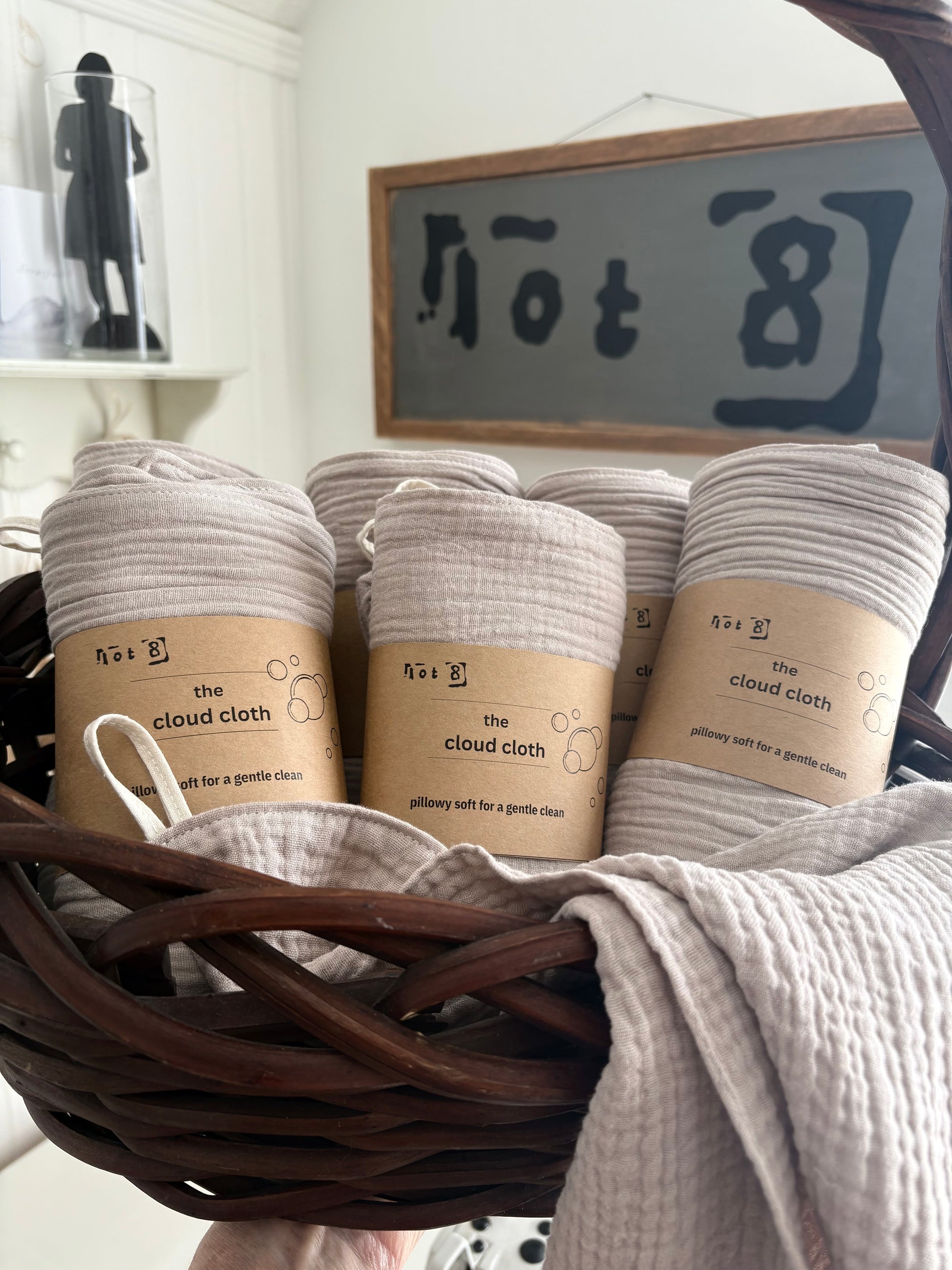 Several rolled fawn-coloured cleansing cloths with kraft paper labels reading 'the cloud cloth' are arranged inside a dark brown wicker basket. A folded matching cloth drapes over the front of the basket.