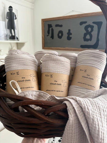 Several rolled fawn-coloured cleansing cloths with kraft paper labels reading 'the cloud cloth' are arranged inside a dark brown wicker basket. A folded matching cloth drapes over the front of the basket.