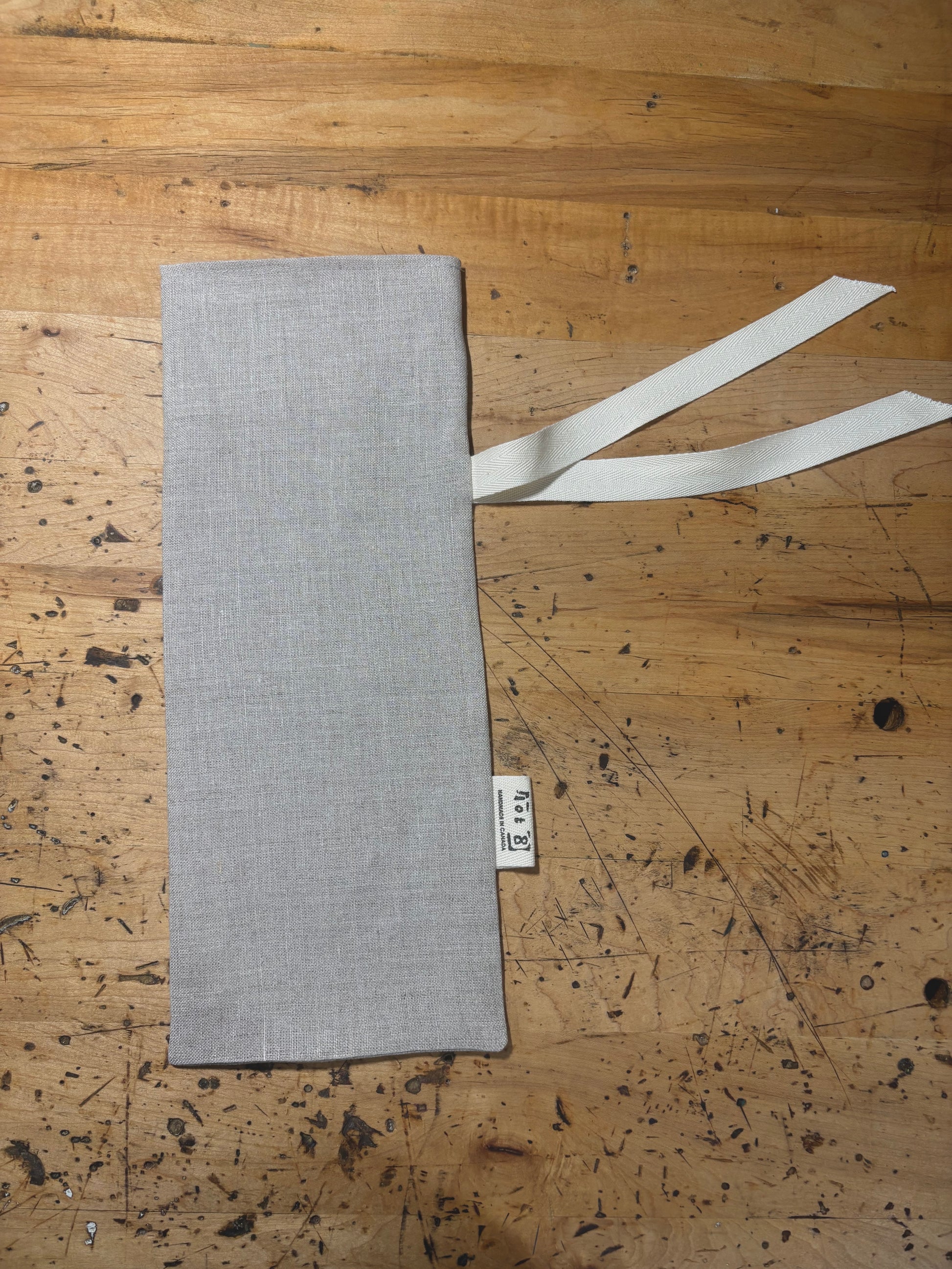 A natural coloured linen wine sack laid flat on a rustic wooden workbench, featuring a cream coloured ribbon tie and a small white fabric brand tag.