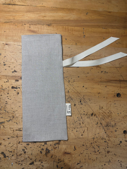 A natural coloured linen wine sack laid flat on a rustic wooden workbench, featuring a cream coloured ribbon tie and a small white fabric brand tag.