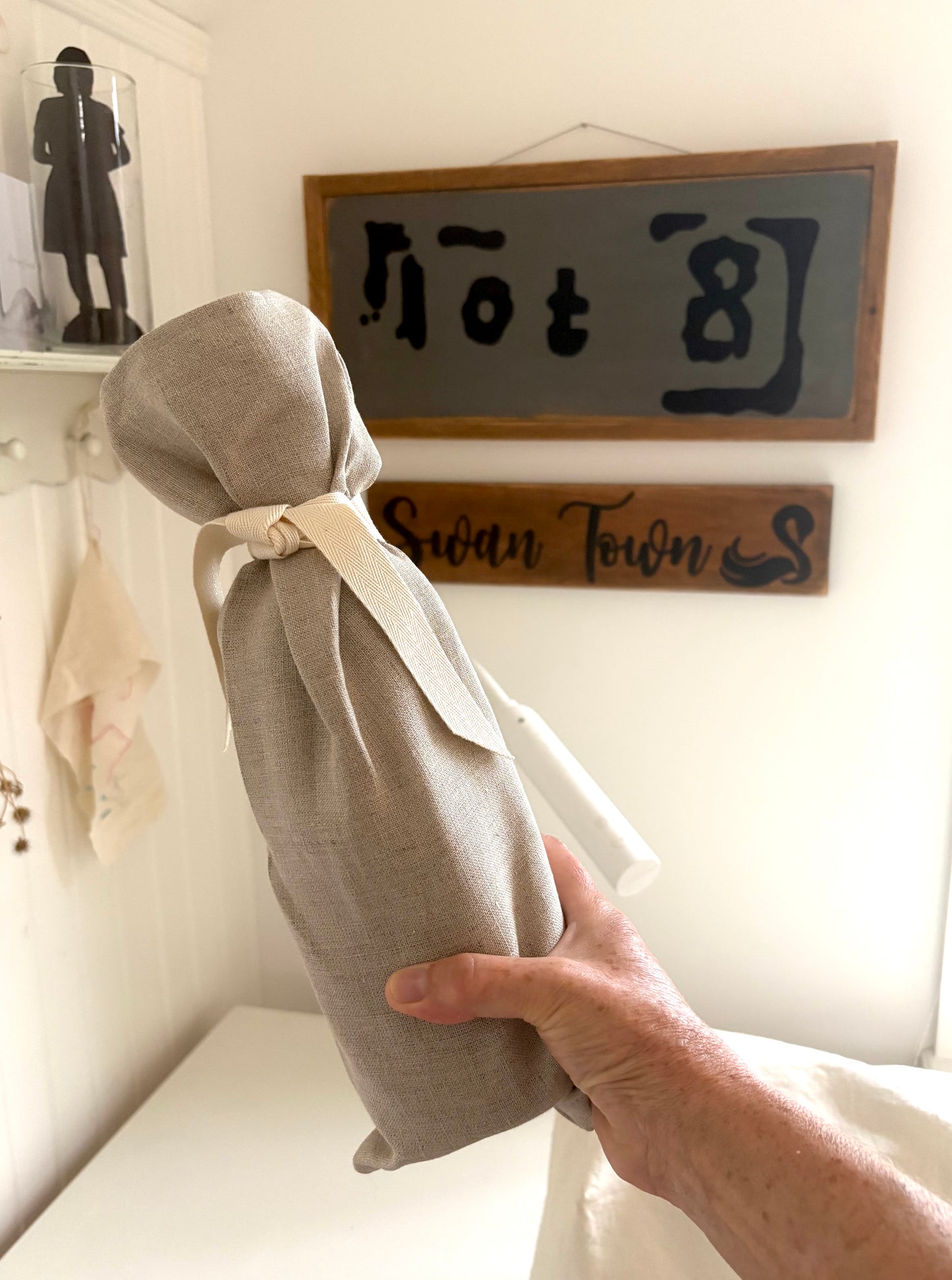 A hand holding a light beige linen fabric pouch tied with a cream ribbon, with a white drawstring visible at the top, in a home interior setting with wooden signs on the wall.