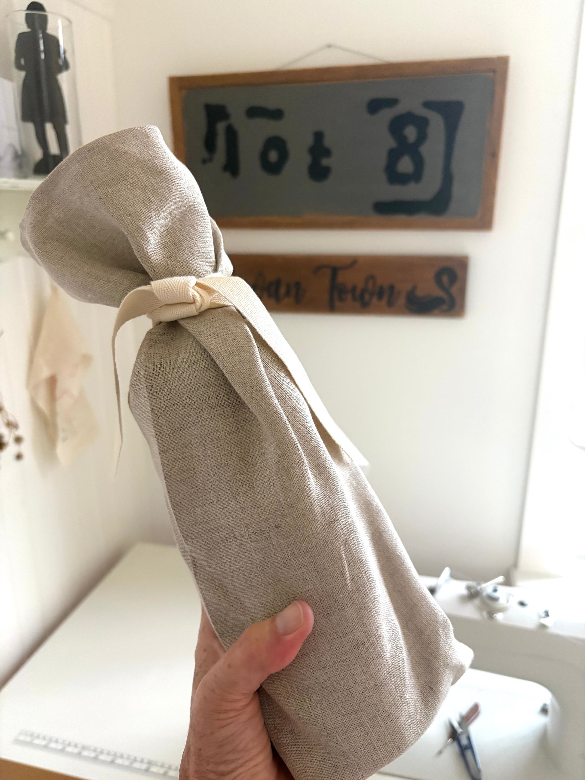 A hand holding a wine bottle encased in a natural linen fabric tote tied with a cream ribbon, set against a softly blurred interior background with wooden signs and a sewing machine.