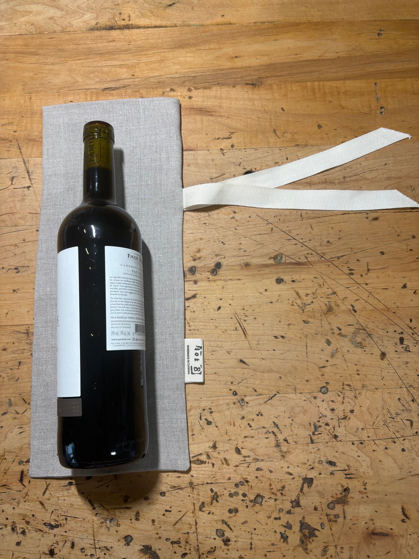 A dark wine bottle resting on a natural coloured linen sack with a white cotton twill ribbon, placed on a scratched wooden tabletop.