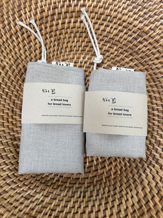 Two light gray linen bread bags with white drawstring cords and paper labels, displayed on a woven wicker surface.