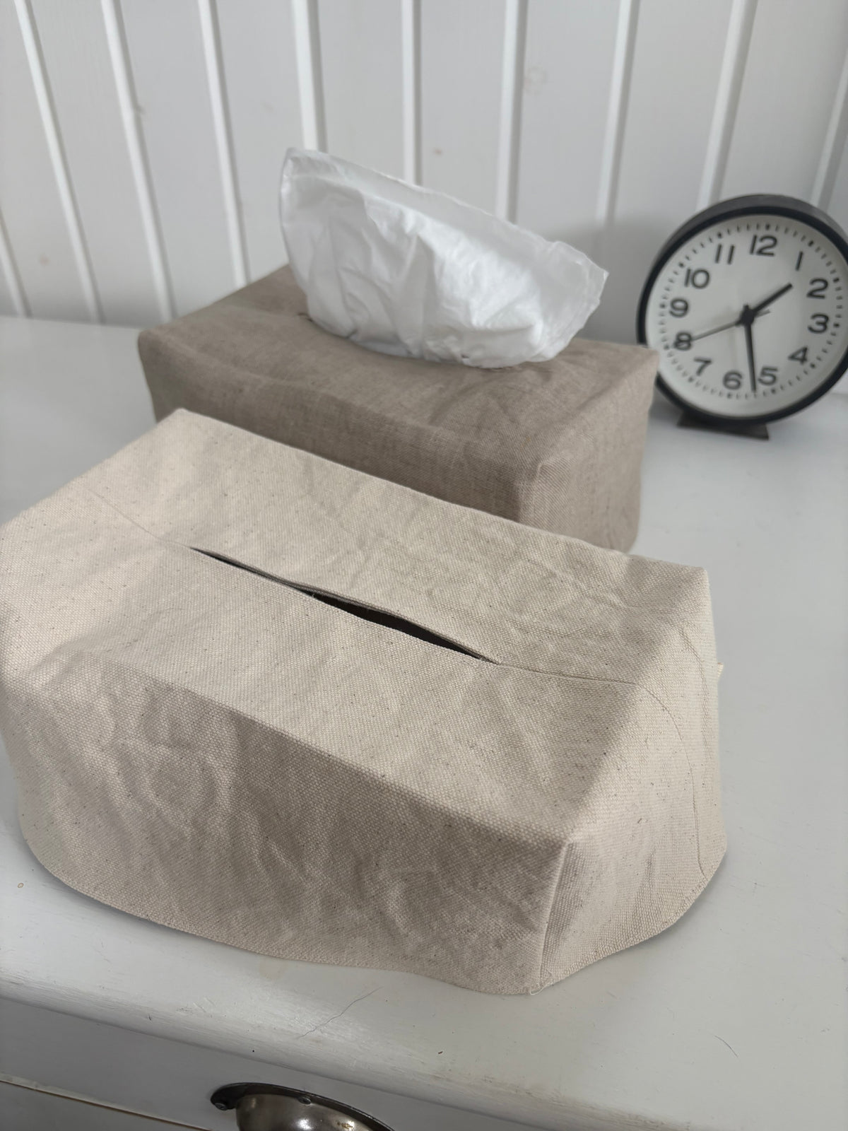Two linen tissue box covers in neutral beige and taupe tones on a white table, with one cover displaying a white tissue and a black analog clock in the background.