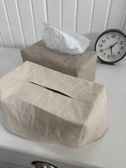 Two linen tissue box covers in neutral beige and taupe tones on a white table, with one cover displaying a white tissue and a black analog clock in the background.