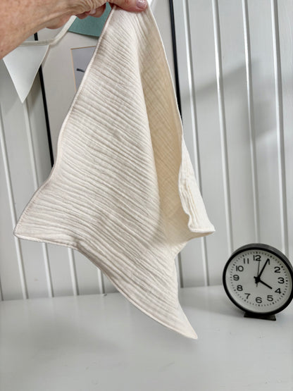 A woman holds a cloud cloth in natural against a wall. A black and white analog clock is in the background.