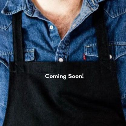 Front view of The Generous Apron in heavy-duty black cotton twill worn by a chef in a modern kitchen setup.  Coming Soon is written across the photo
