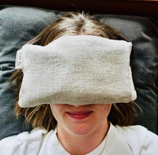 A woman resting with a linen eye pillow over her eyes. She has a slight smile as she relaxes.