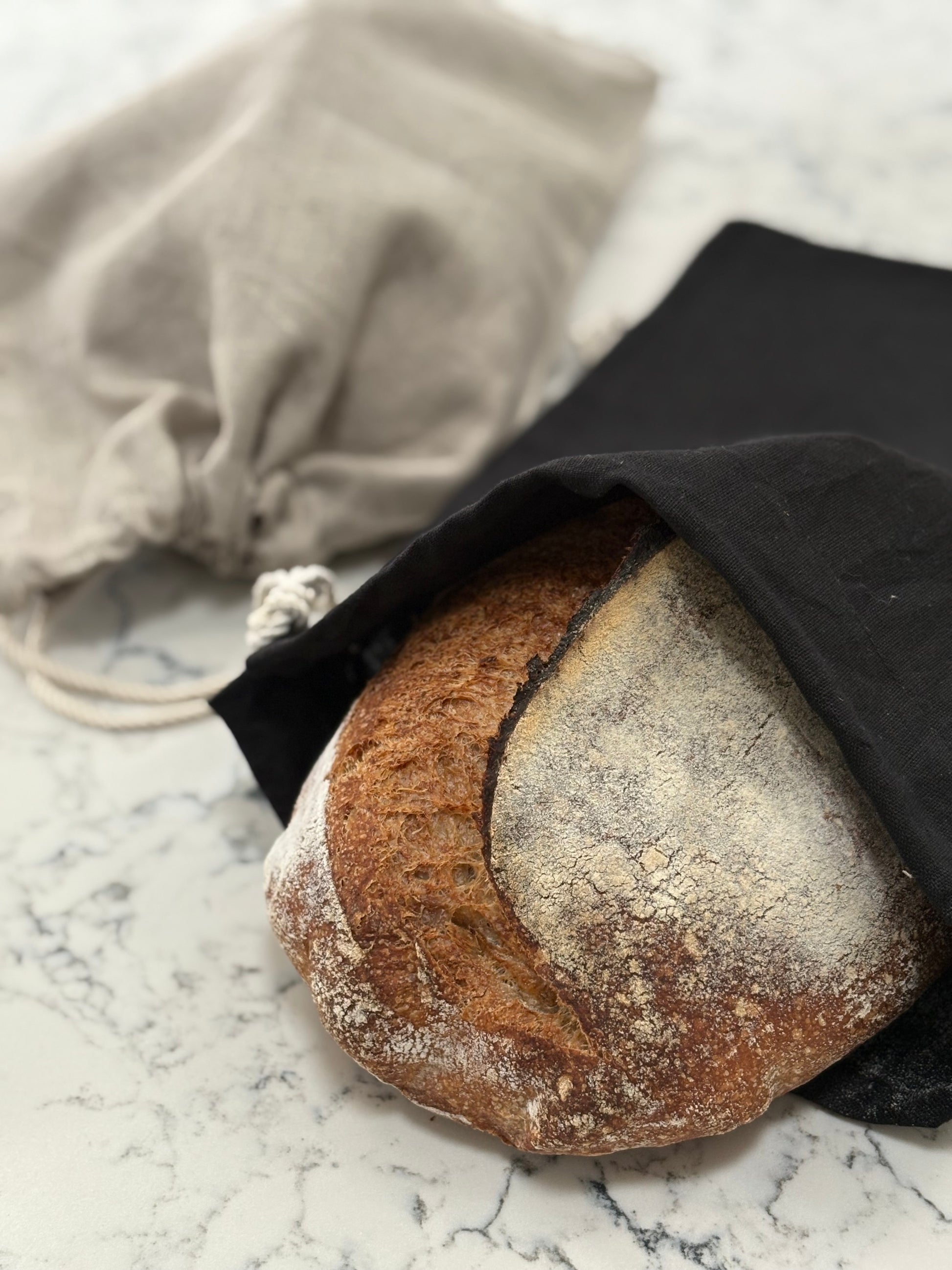 Loaf of bread pokes out of a hand-sewn  black linen bread bag  on a marble surface with a natural linen bread bag  in the background