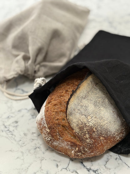 Loaf of bread pokes out of a hand-sewn  black linen bread bag  on a marble surface with a natural linen bread bag  in the background
