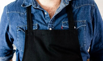 Front view of The Generous Apron in heavy-duty black cotton twill worn by a chef in a modern kitchen setup.