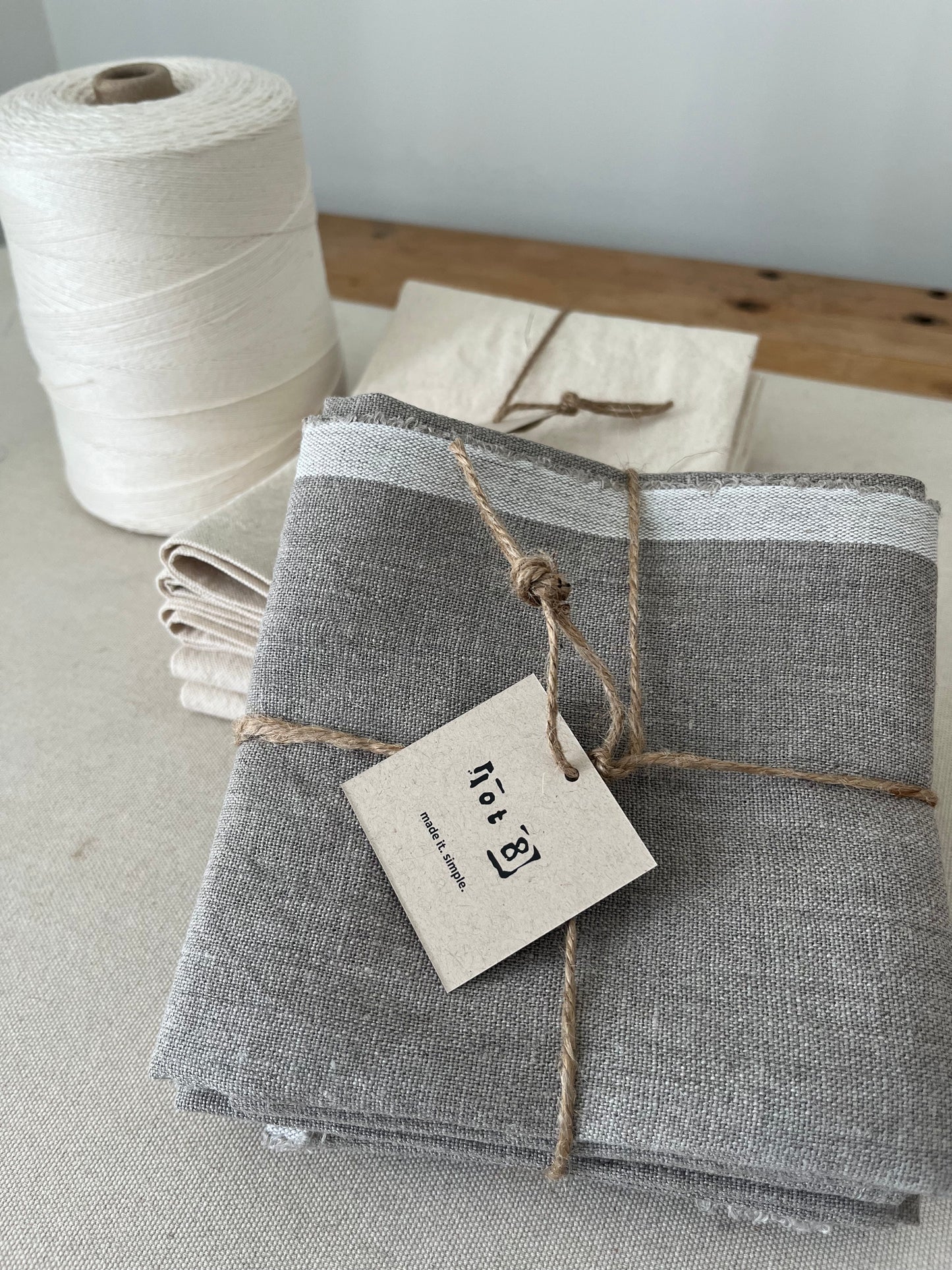 A set of 4 linen napkins is tied with a sisal string with a lot8 kraft paper tag. It sits in front of a set of canvas napkins also tied. In the background sits a spool of twine.
