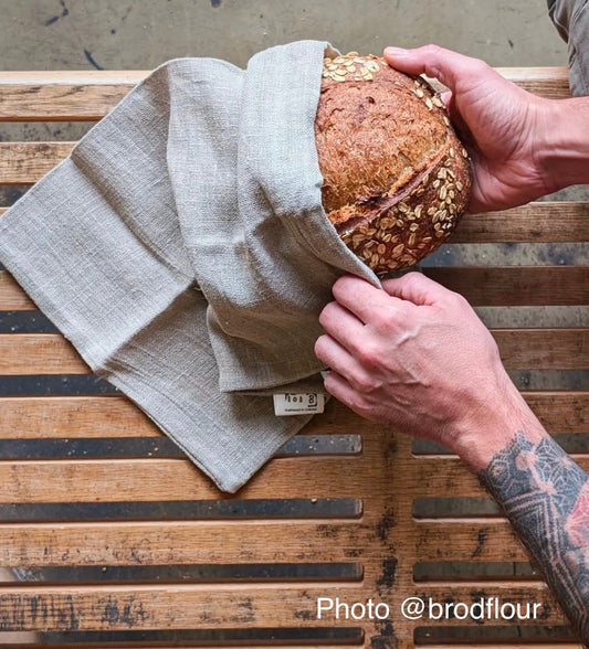 Hand-sewn natural linen bread bag holds a bakery style loaf studded with oatmeal.  A tattooed arm holds the bag.