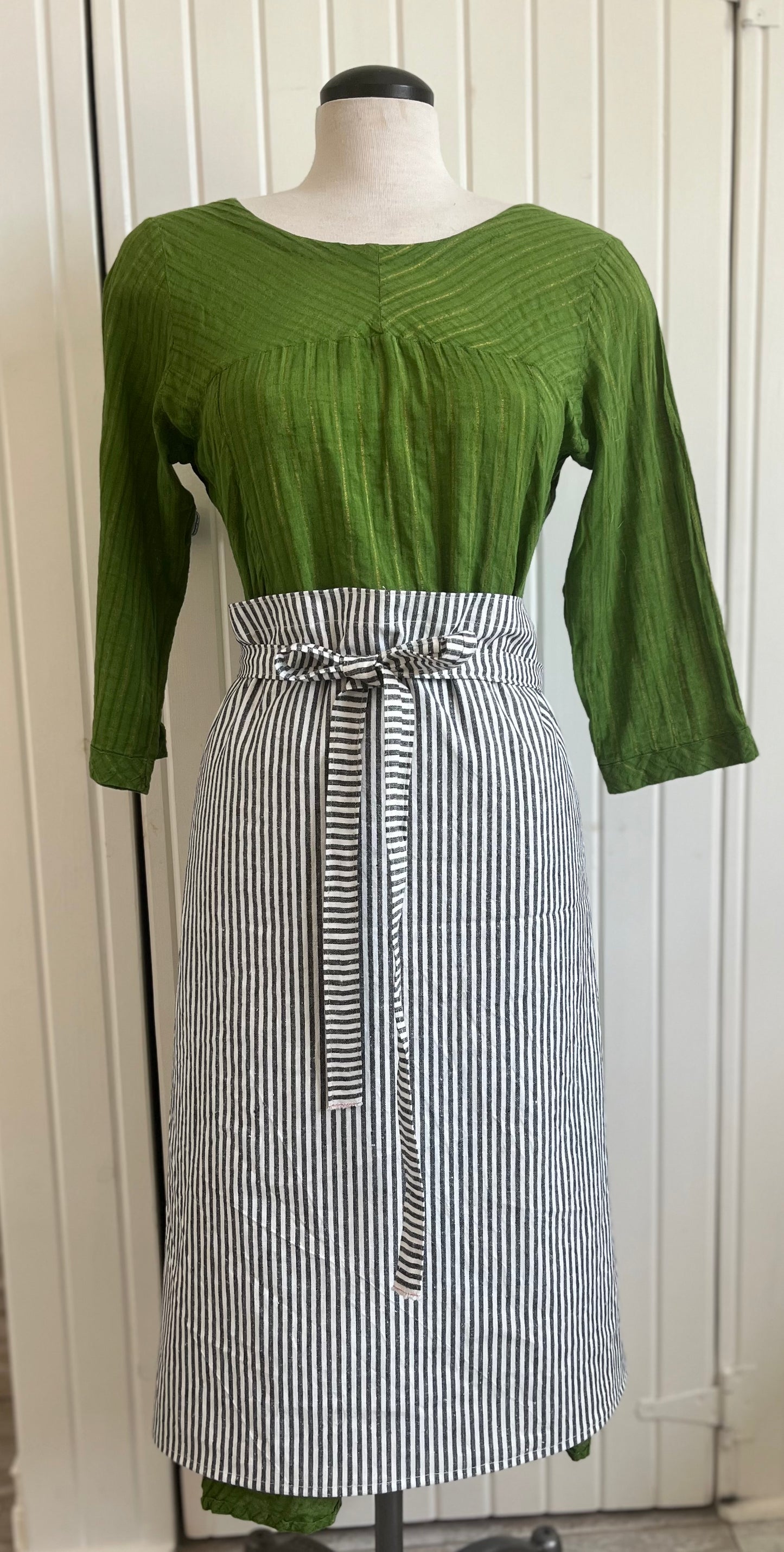 A dressmaker's Judy dressed in an apple-green dress sports and black and white striped half apron. The bow is tied in the front. She looks very elegant.