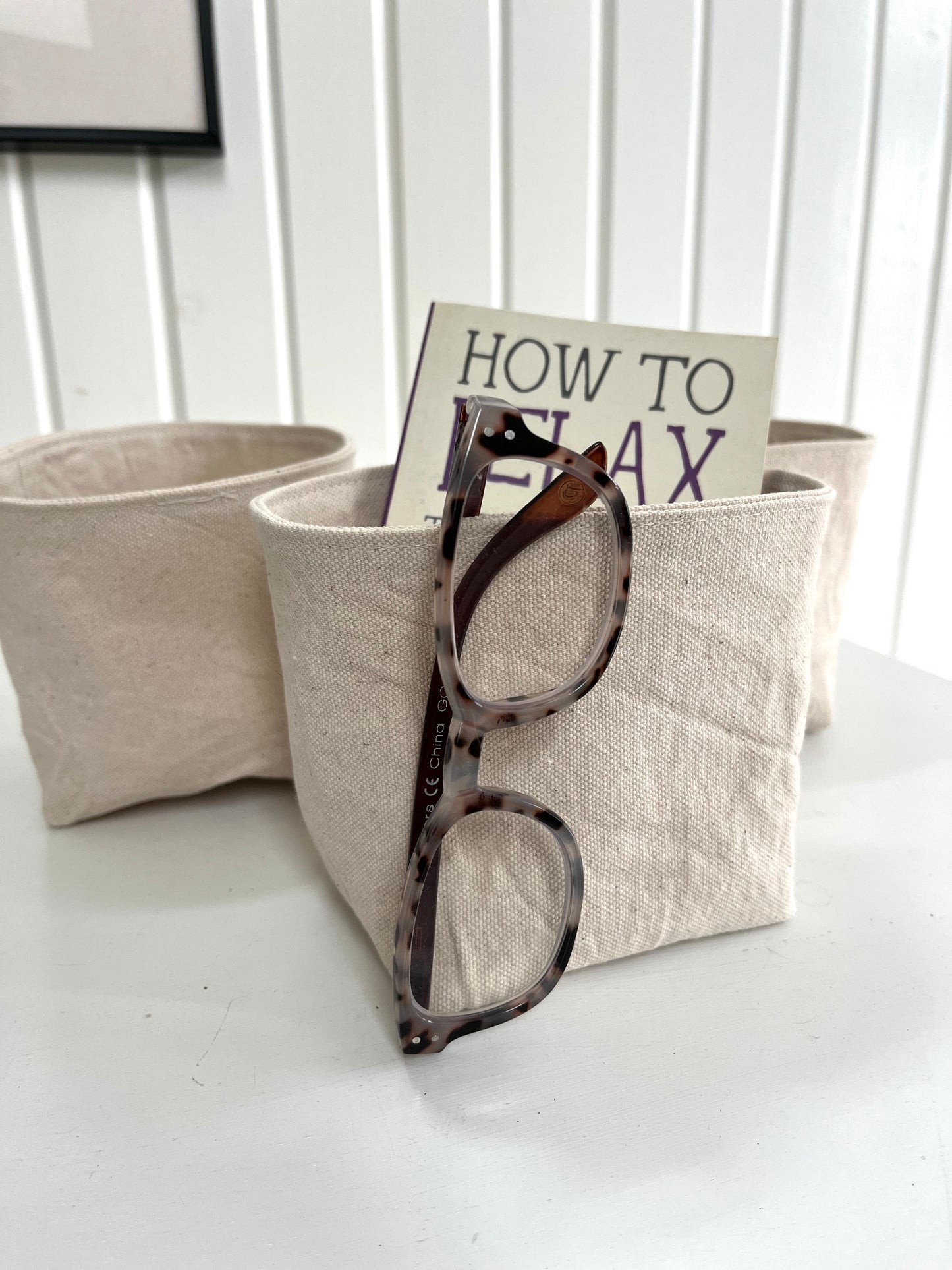 The small bucket sits on a white dresser. Inside it is a small book titled "How to Relax". A pair of reading glasses hangs from the front of the bucket. Two additional buckets sit in the background.