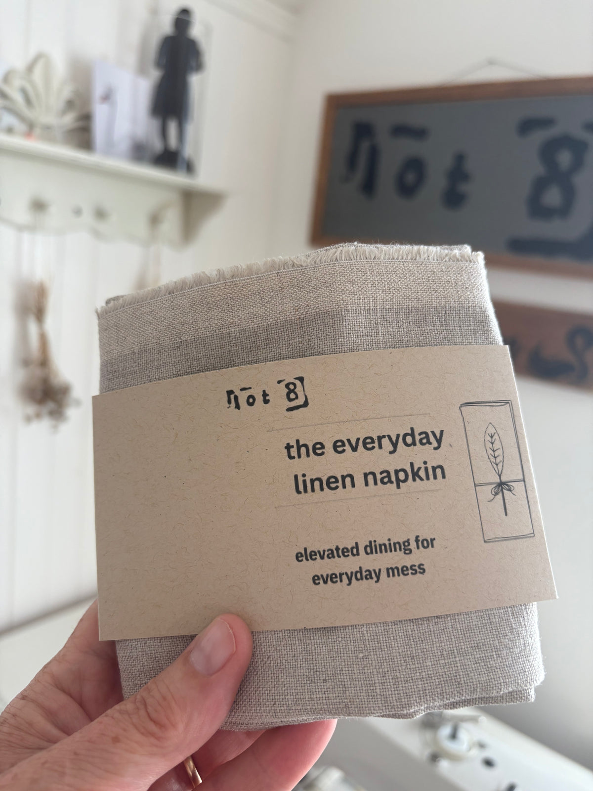 A hand holding a folded linen napkin with a kraft paper label that reads 'the everyday linen napkin' in a home setting.