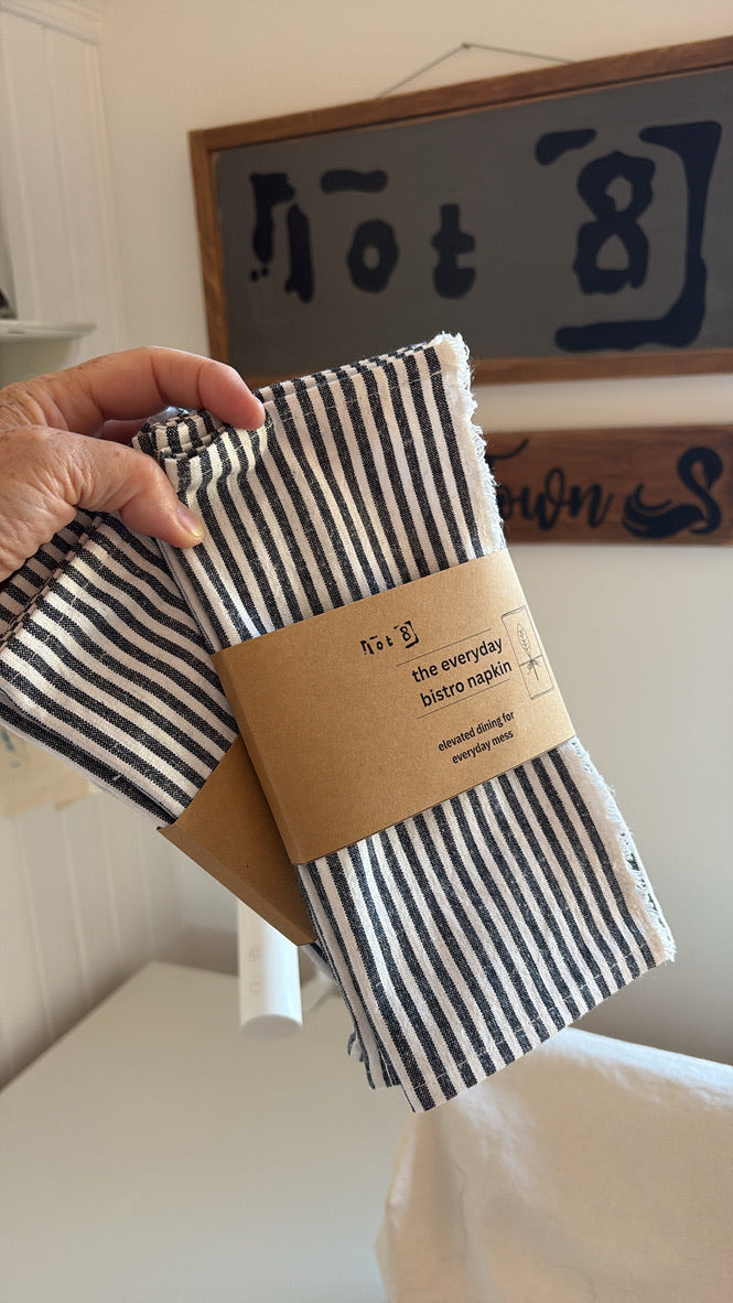 The set of striped napkins is wrapped with a kraft paper belly band labelled 'the everyday bistro napkin". The product description and care instructions are on the back of the packaging.

