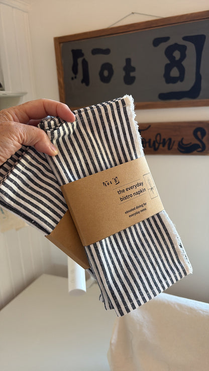 The set of striped napkins is wrapped with a kraft paper belly band labelled 'the everyday bistro napkin". The product description and care instructions are on the back of the packaging.
