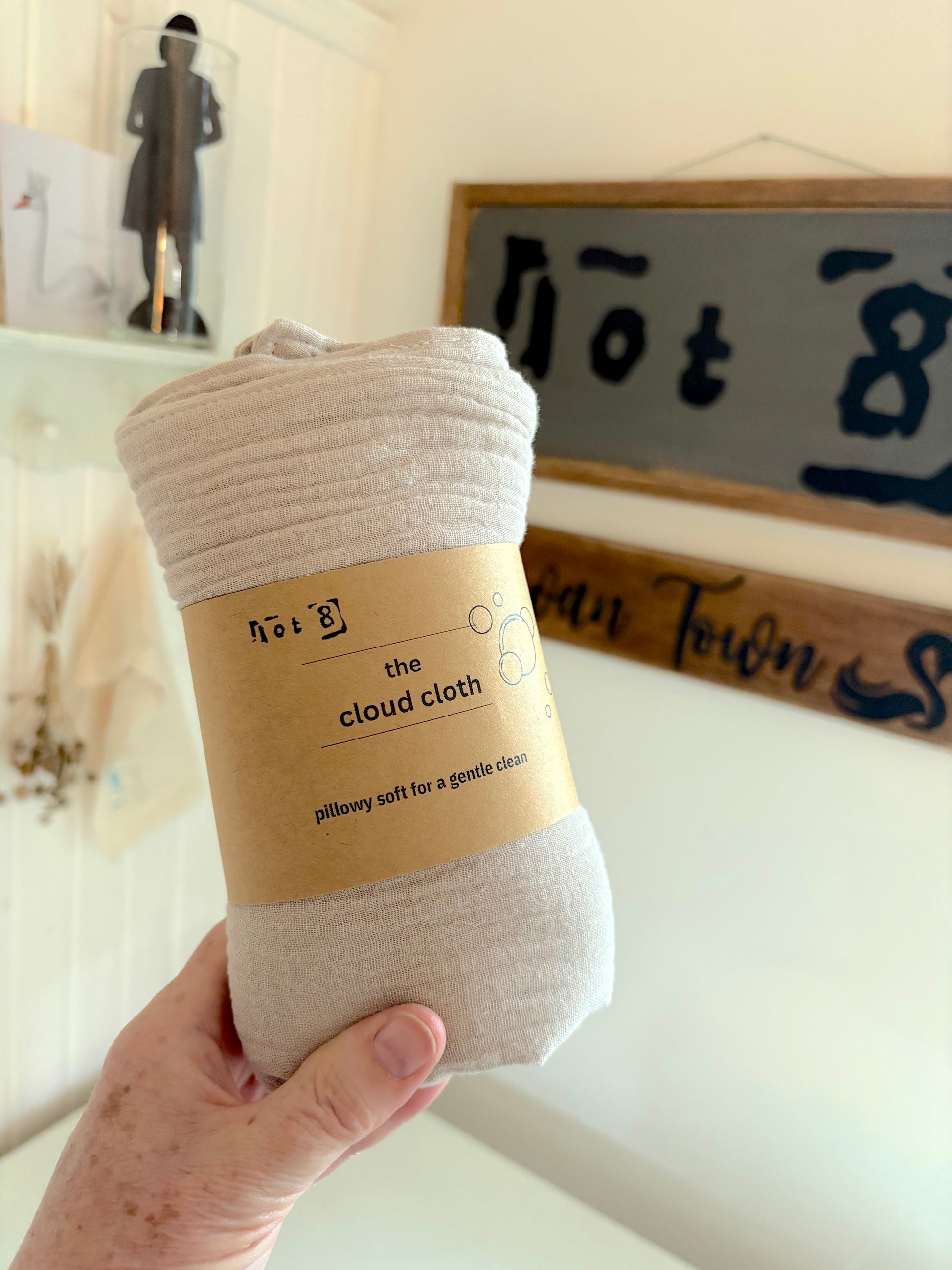 A hand holds a rolled set of 4 fawn-coloured cleansing cloths with a kraft paper label that reads 'the cloud cloth' and 'pillow soft for a gentle clean.' The background shows a softly blurred home interior with a framed sign and shelf decor.