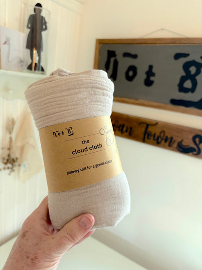 A hand holds a rolled set of 4 fawn-coloured cleansing cloths with a kraft paper label that reads 'the cloud cloth' and 'pillow soft for a gentle clean.' The background shows a softly blurred home interior with a framed sign and shelf decor.