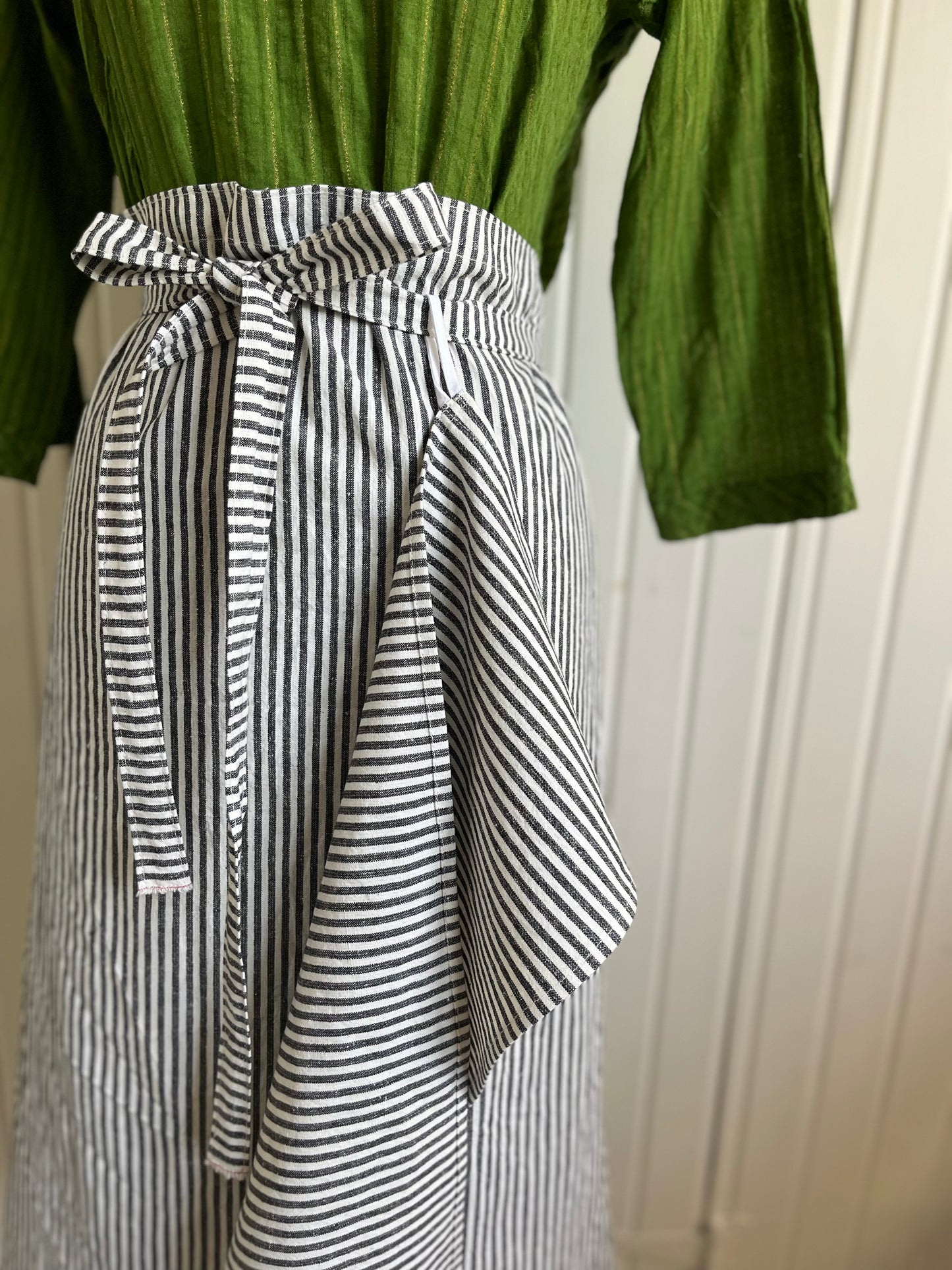 A close up of the black and white striped bistro apron on the dressmaker's Judy. The waist strap of the apron is threaded through the loop of a coordinating hand-towel .