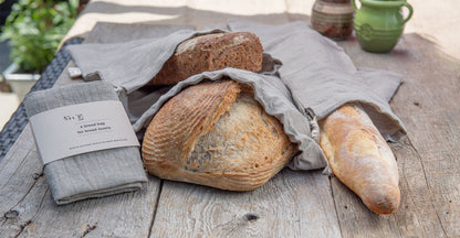 Various loaves of bread nestle in hand-sewn linen bread bags on a rustic wooden table. A packaged bread bag with the label "A Bread Bag for Bread Lovers" sits off to the side.