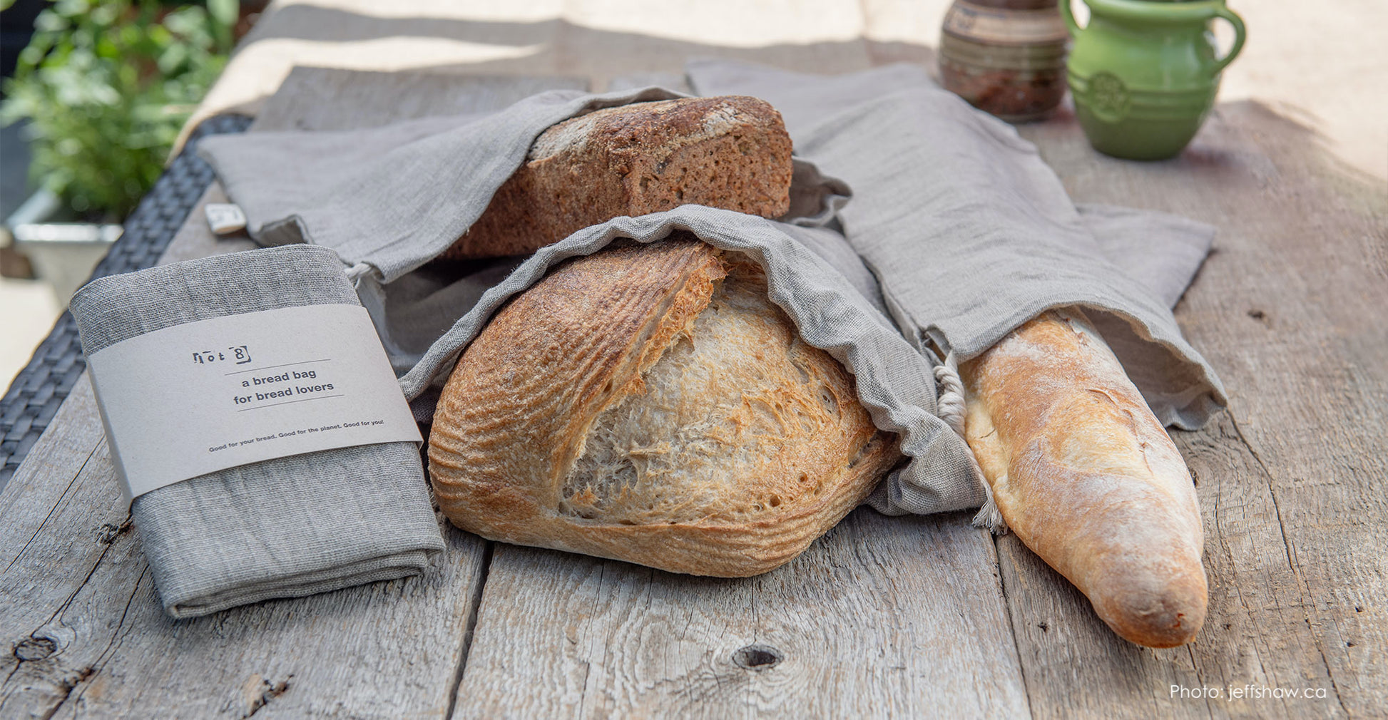 Rustic loaves in 3 sizes are nestled into linen bread bags and lay on top of a rustic wooden table top. One packaged bread bag sits in the foreground