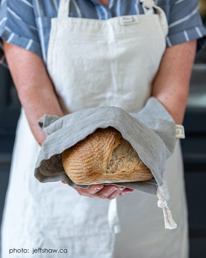 A woman wearing a canvas apron extends her arms holding a loaf of bread in a hand-sewn linen bread bag. 