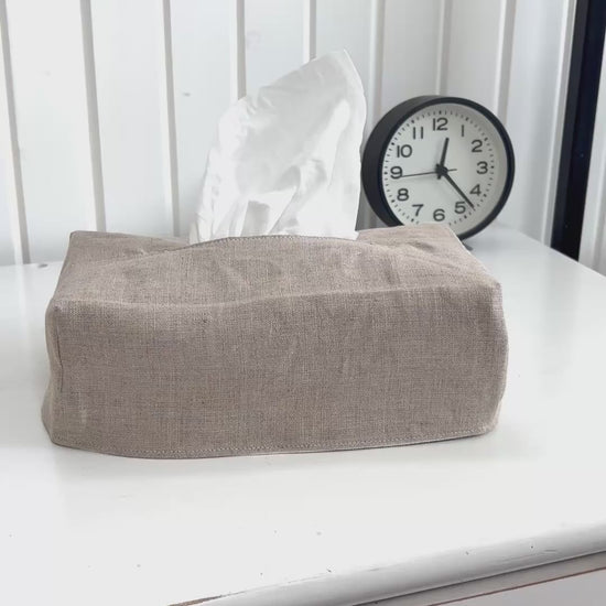 The tissue box cover in white canvas sits on a white dresser. A woman removes the cover, reverses it and covers the tissue box again, this time revealing a natural linen.