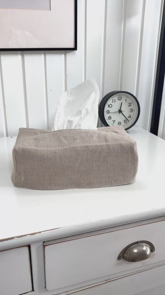 The tissue box cover in white canvas sits on a white dresser. A woman removes the cover, reverses it and covers the tissue box again, this time revealing a natural linen.