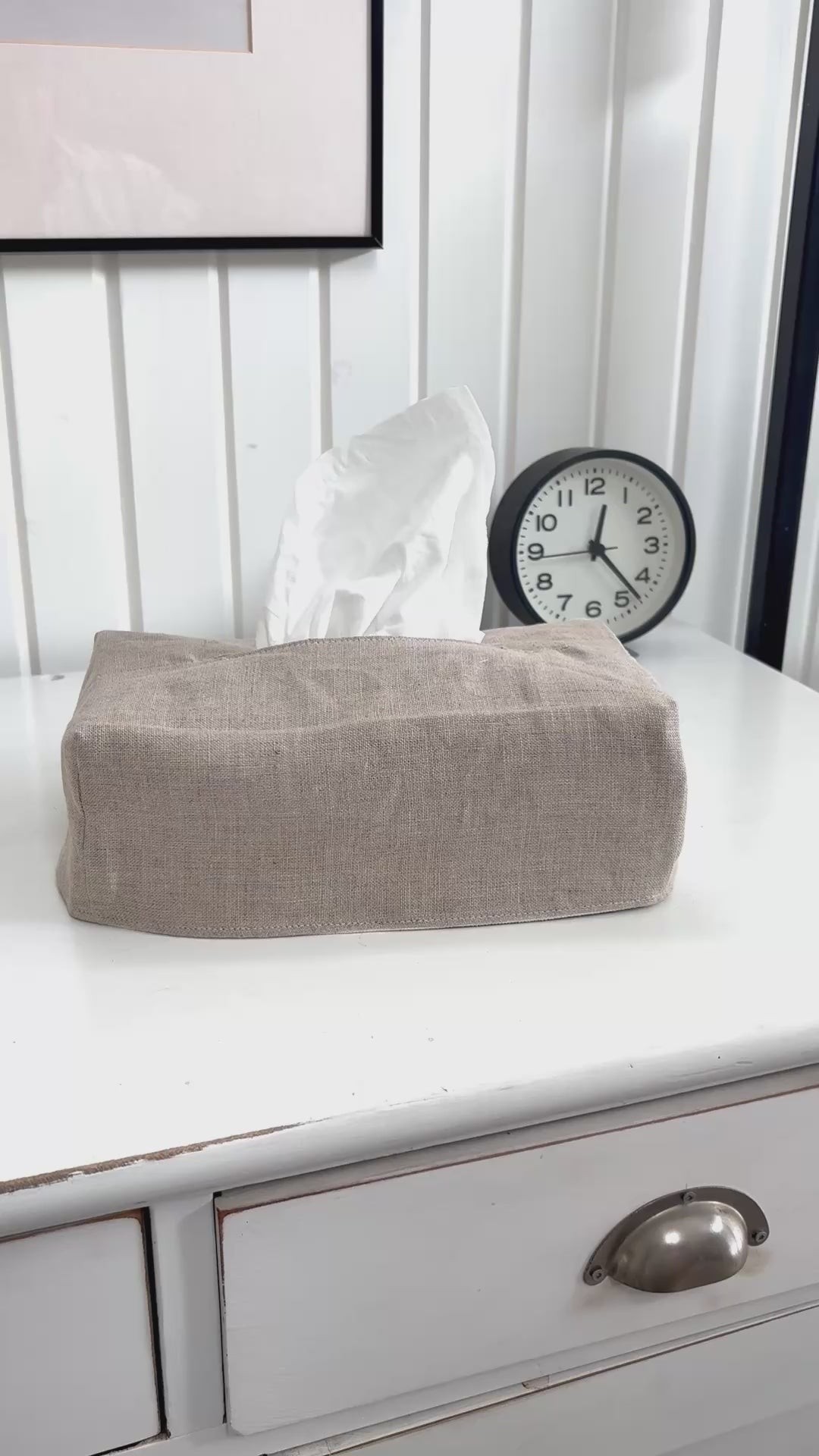 The tissue box cover in white canvas sits on a white dresser. A woman removes the cover, reverses it and covers the tissue box again, this time revealing a natural linen.