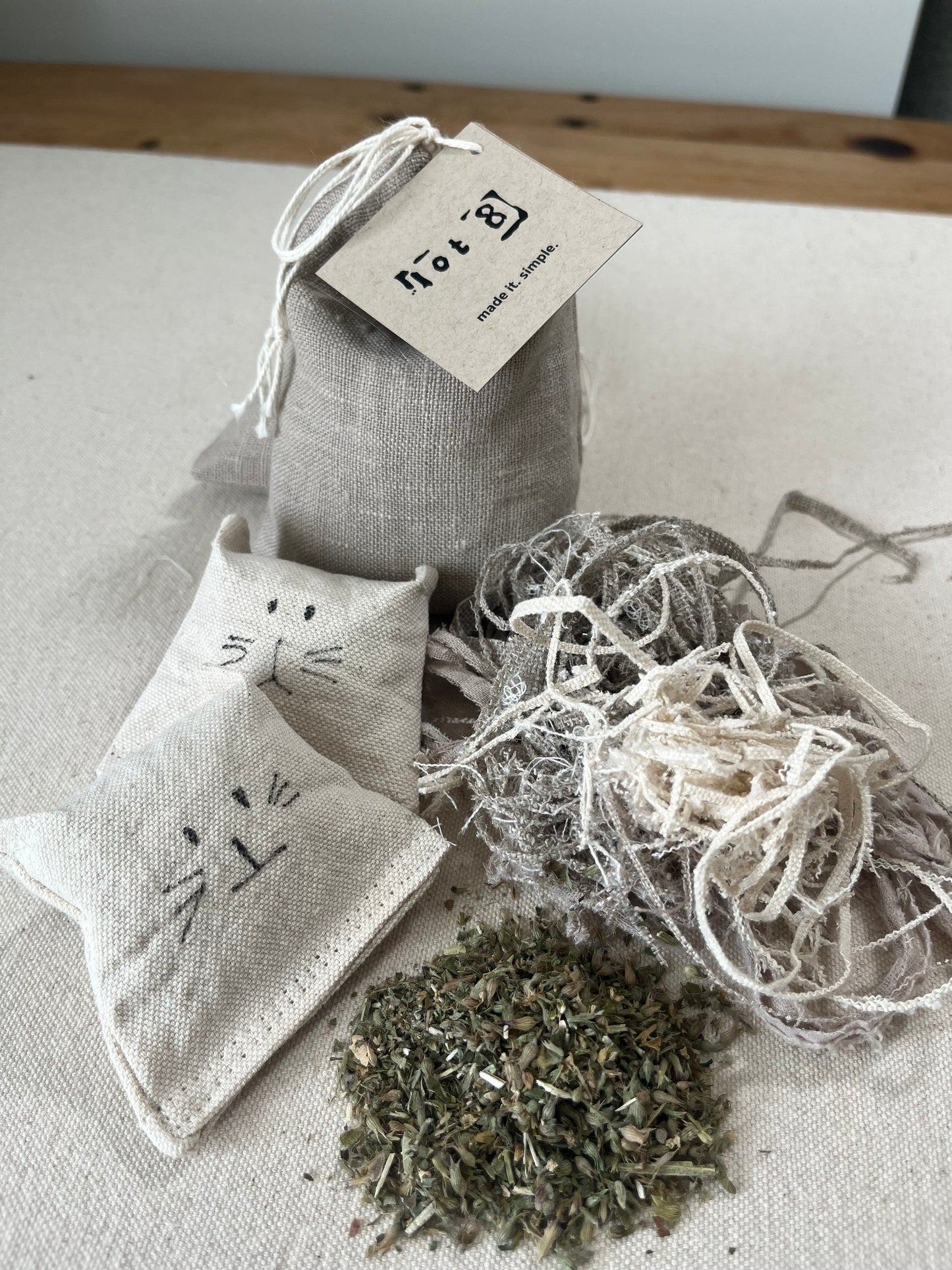 2 natural cat toys with adorable faces lay on a table. Beside them are small piles of their filling; a pile of organic cat nip and a pile of natural fabric scraps. Behind them sits a linen pouch filled with cat grass seeds.