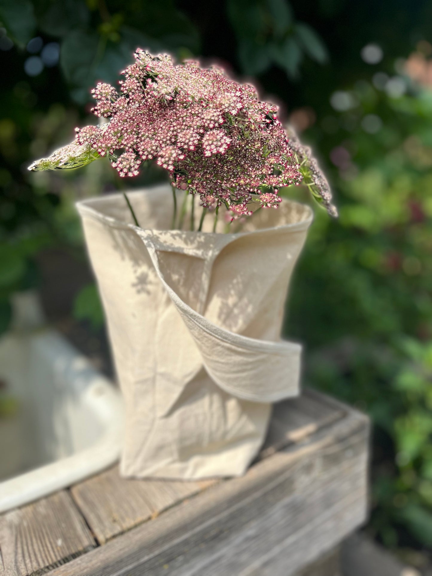 A single handed cotton canvas tote sits on a rustic table top. Pink and purple blooms stick out the top of the tote.