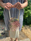 A woman in a blue dress holds a cotton canvas one-handled tote. Pink and purple blooms pop out the top. A garden is blurred in the background.