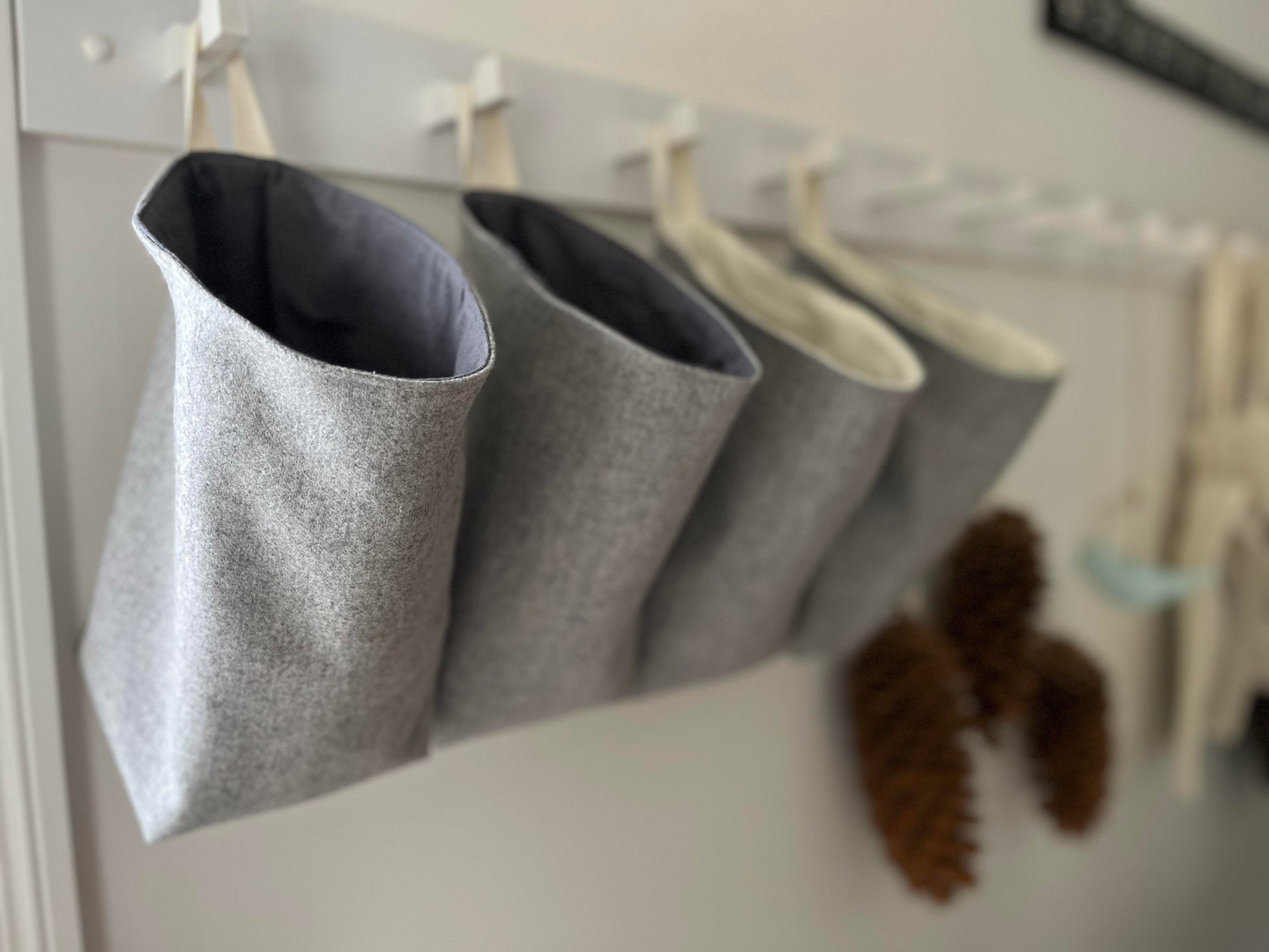 4 grey all purpose sacks hang from a white hanging rail in a foyer. A couple of pinecones are blurred in the distance hanging from the same rail.