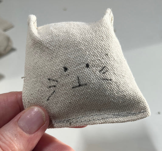 A close up of the all natural cat toy. It is a canvas square with little ears sewn into the top of it. A simple cat face is drawn onto the front of the square. it's adorable.