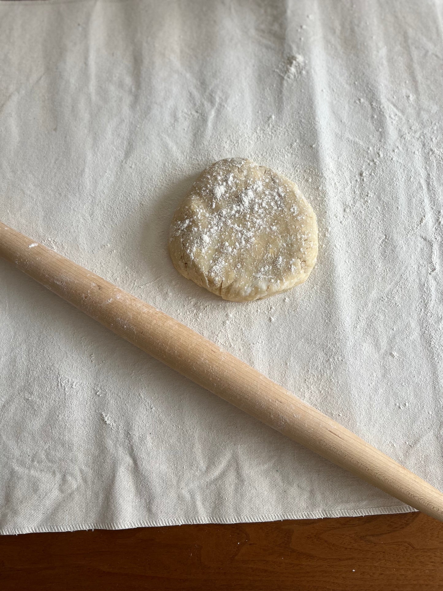 A small disc of dough sits on a pastry cloth. Rolling pin in the foreground; jar of flour in the background