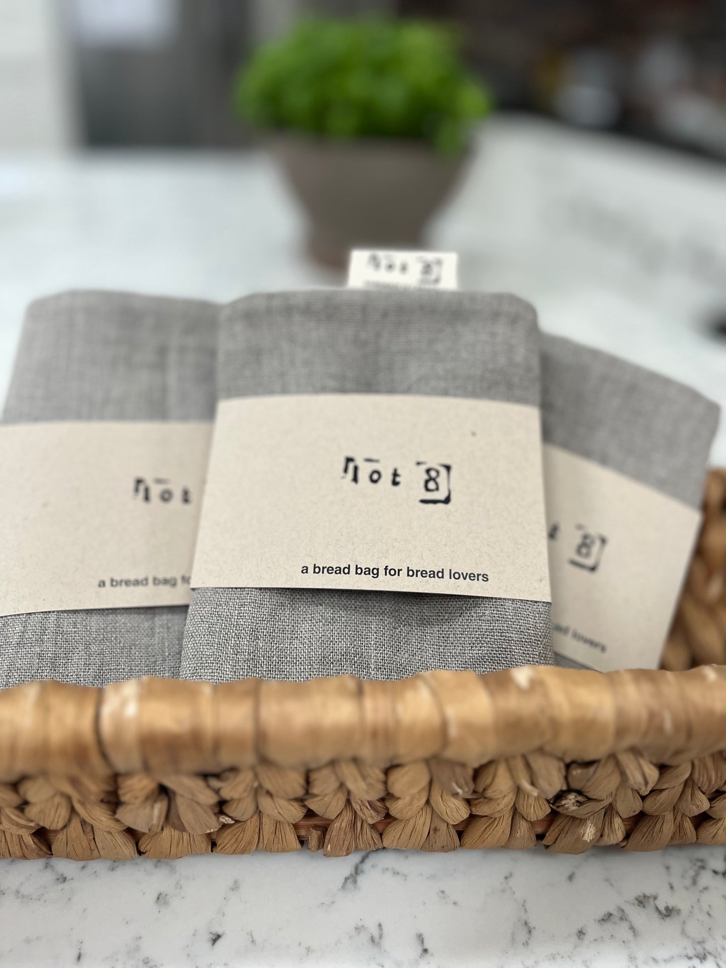 3 linen bread bags are nestled in a woven basket (not included). Each is wrapped in a lot8 kraft paper belly band with 'a bread bag for bread lovers ' printed on the front.A pot of basil is blurred in the background.