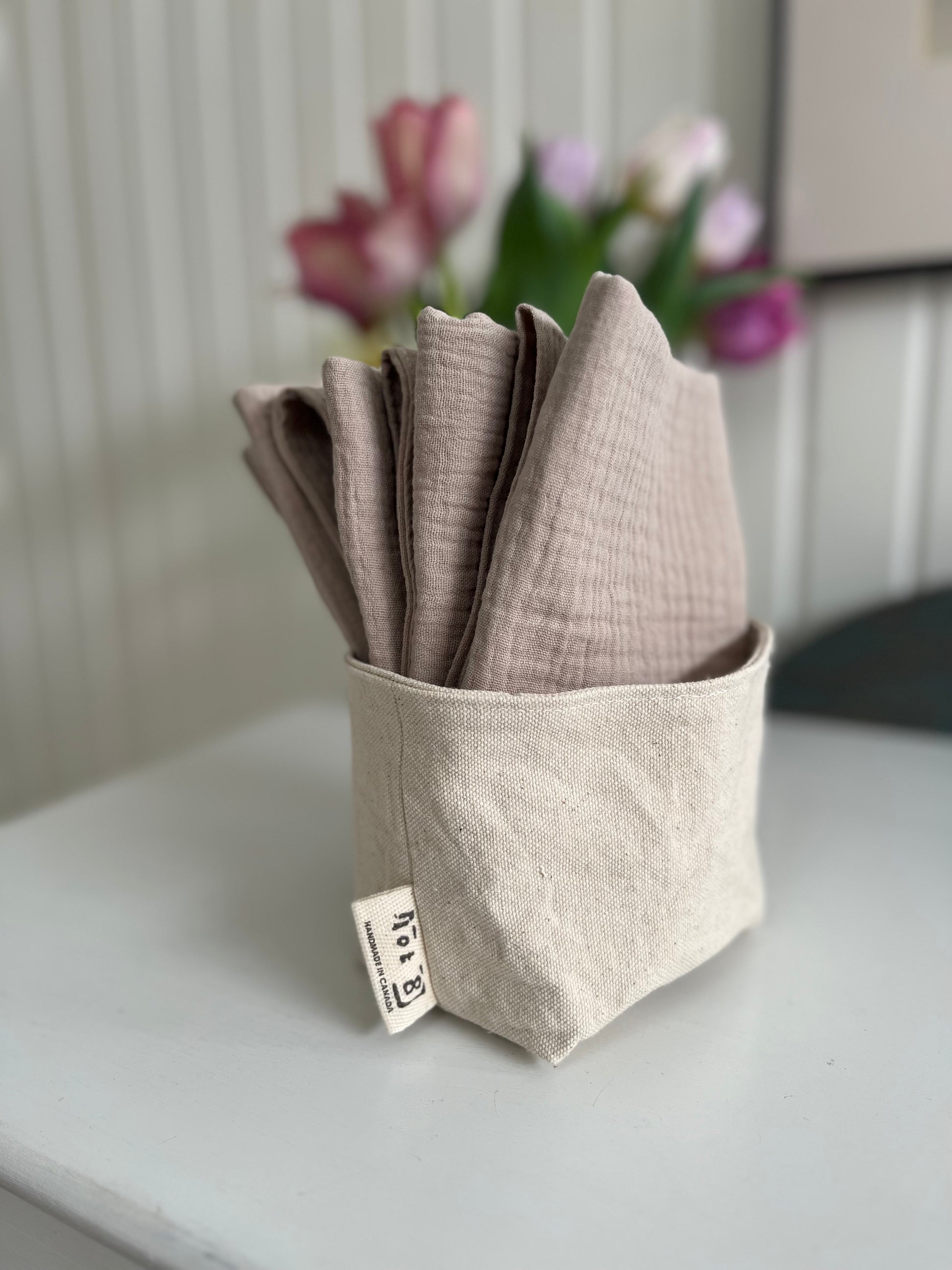 4 organic cotton facial cleansing cloths in 'faun' are nestled in a fabric organizer called The Bucket. A vase of pink tulips is blurred in the background.