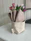 4 organic cotton facial cleansing cloths in 'faun' are nestled in a fabric organizer called The Bucket. A vase of pink tulips is blurred in the background.