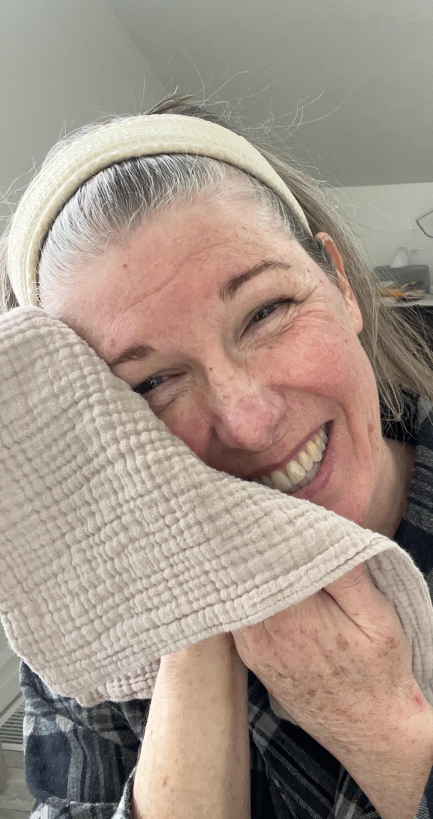 A woman smiles into the camera. Her hair is pulled back with a hair band. She rests the cloth on one side of her face.