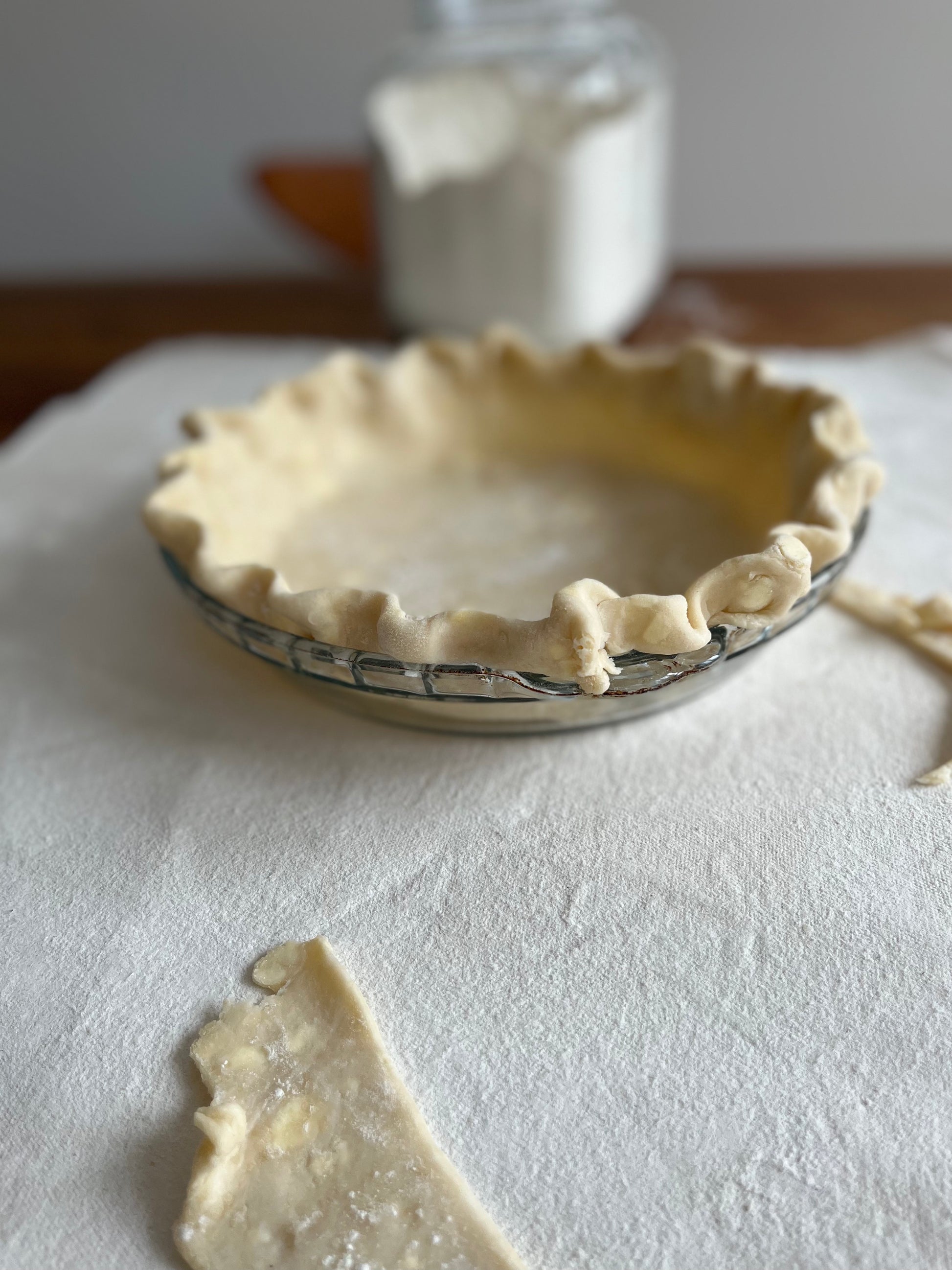 A crimped pie crust in a pie plaste sits atop a pastry cloth. A jar of flour is in the background.