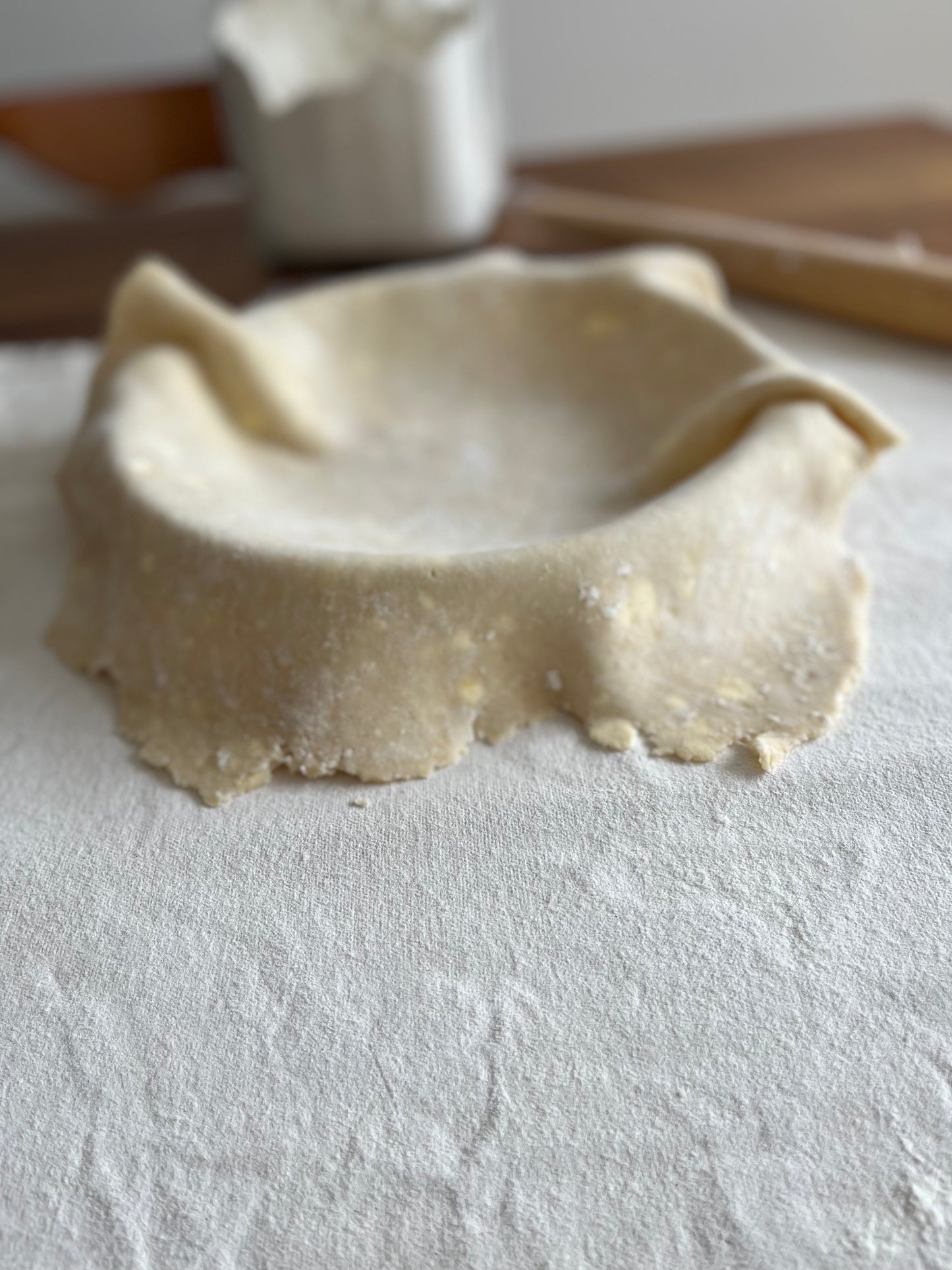 Pie dough is draped in a pie plate. The pie plate sits on a pastry cloth. Rolling pin and jar of flour in the background.