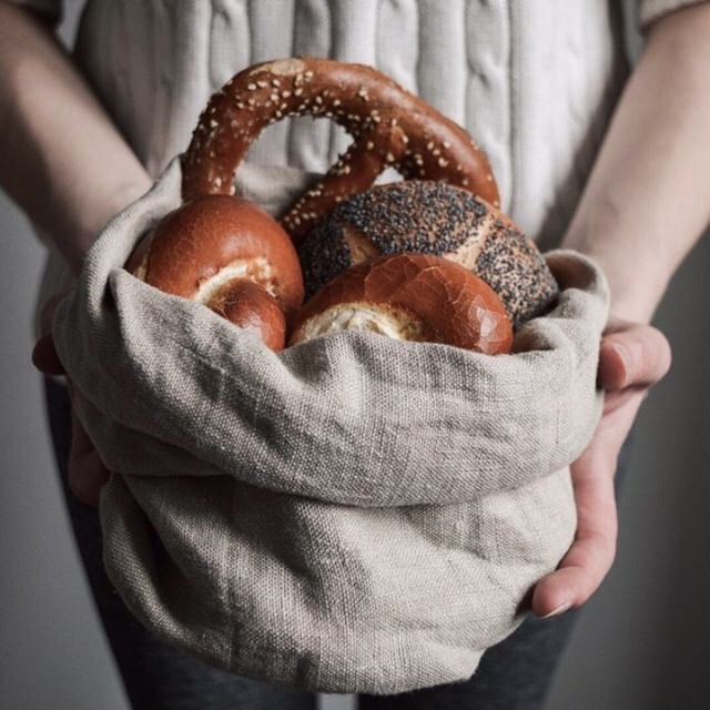 A woman holds a hand-sewn linen bread bag with a variety of rolls peaking from the top of it. 